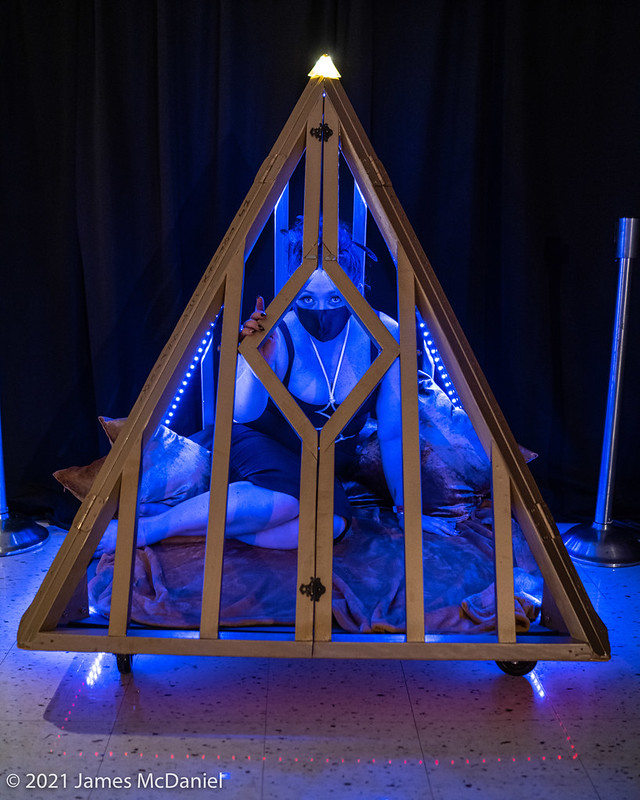 Femme presenting person in a pyramid shaped cage washed in blue light from the 2021 Seattle Erotic Art Festival
