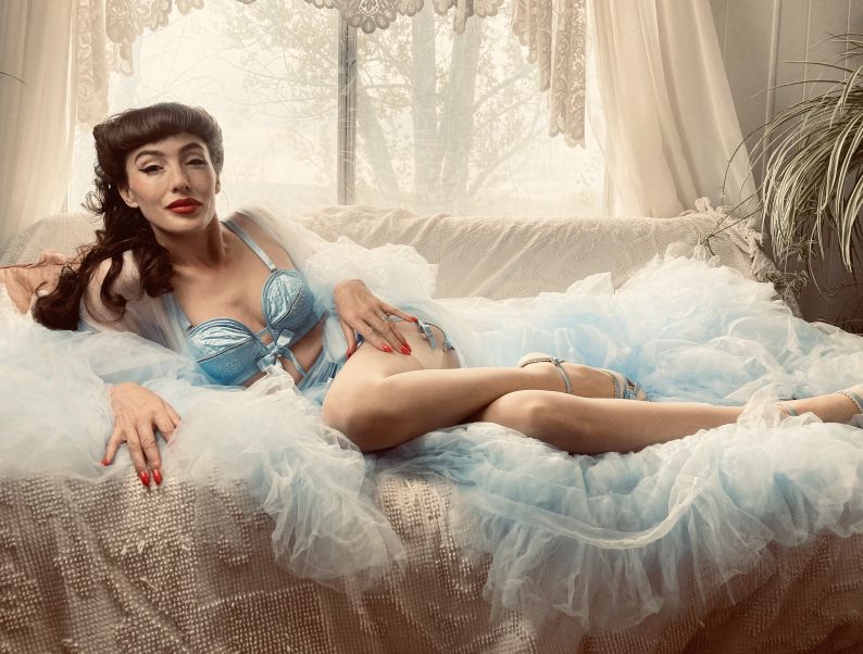 2024 Interactive Experiences: Sugar May reclined on a couch wearing a voluminous dressing gown over blue lingerie.