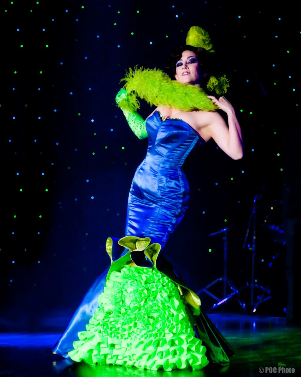 2024 Performance Curators: Miss Indigo Blue on stage mid-performance in a blue and green dress.